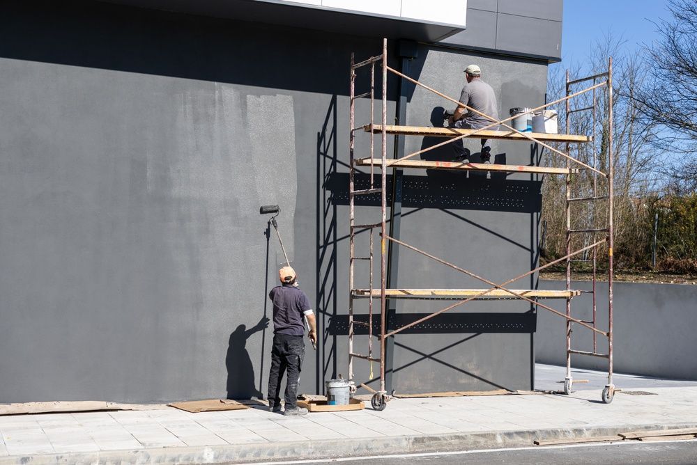 Trabajadores en andamios pintando el exterior de un edificio gris en un día soleado.