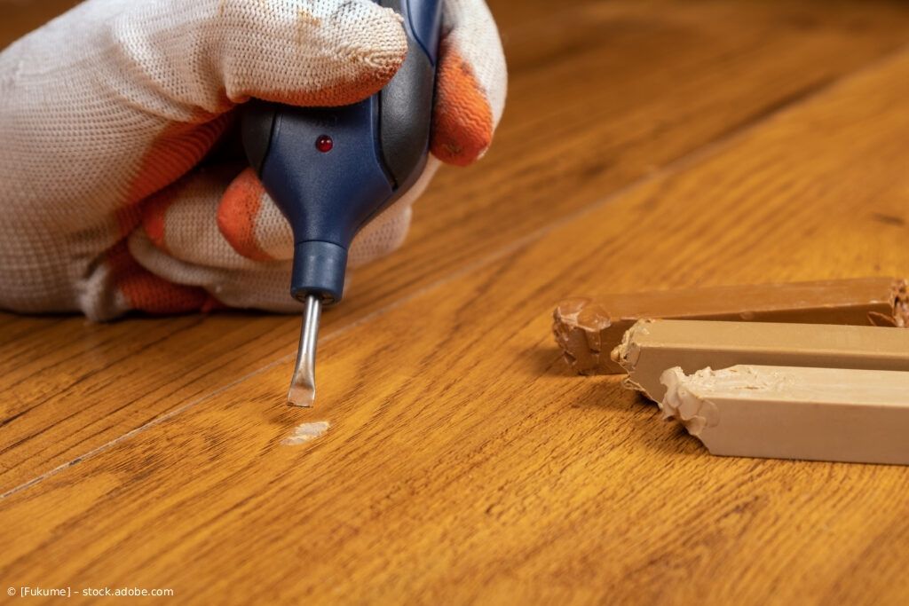 Eine behandschuhte Hand benutzt ein erhitztes Wachsreparaturwerkzeug, um einen Kratzer auf einem Holzboden auszubessern; 