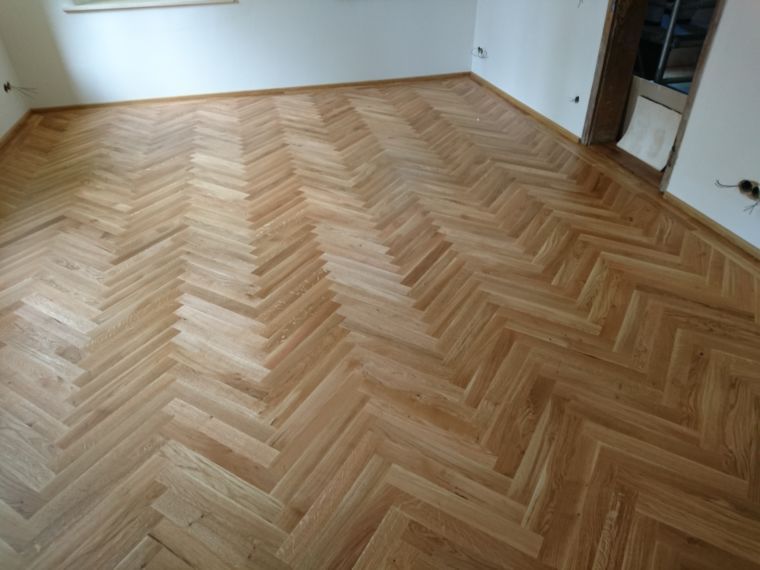 Ein heller Holzboden, verlegt im Fischgrätmuster, in einem leeren, weiß getünchten Raum.