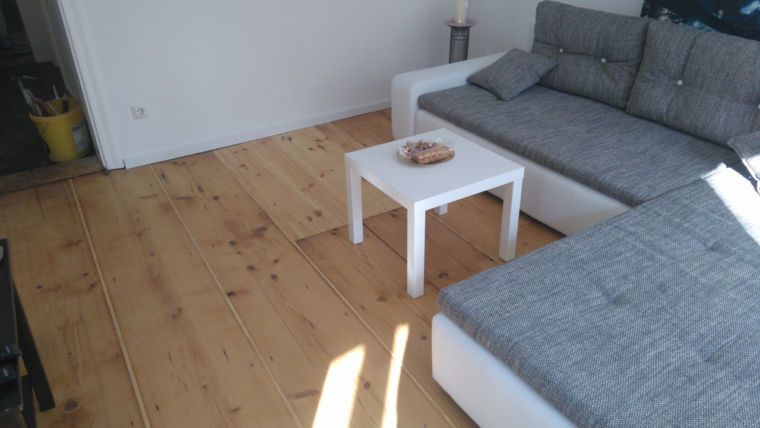 Ein helles Wohnzimmer mit Holzboden, grauem Ecksofa, weißem Couchtisch und weißen Wänden.