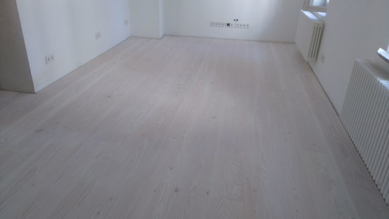 Ein leerer Raum mit hellem Holzboden und weißen Wänden, an dessen rechter Wand sich ein Heizkörper befindet.