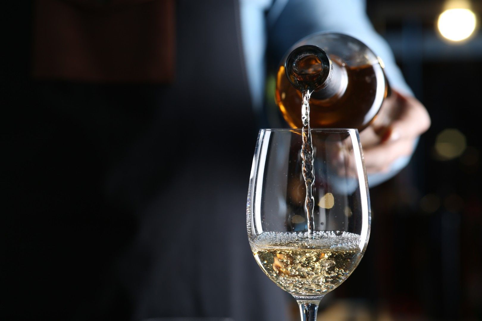 Une main verse du vin ambré dans un verre à pied sur fond sombre.