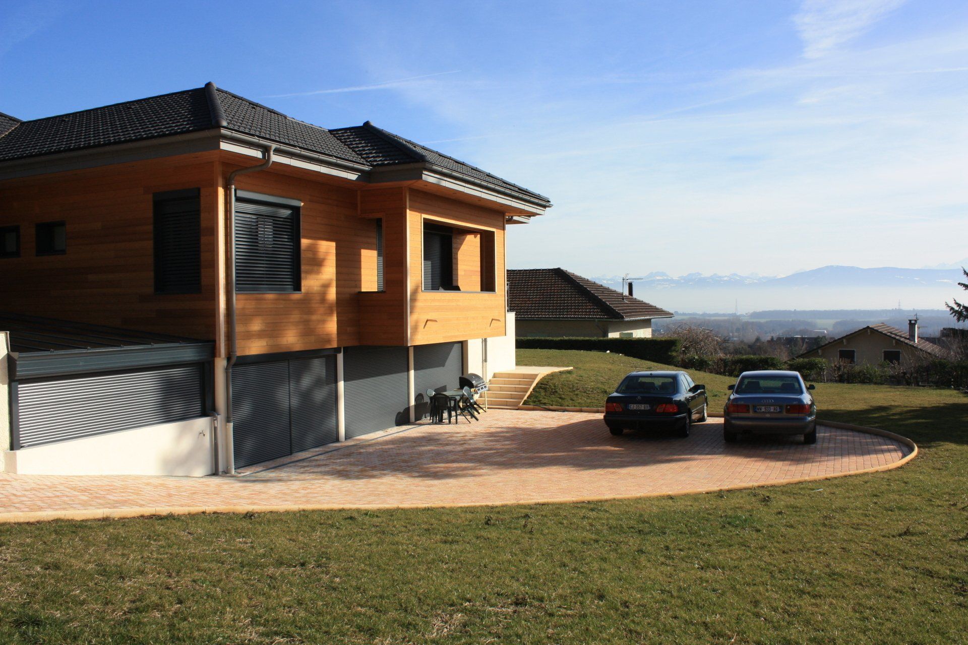 Maison moderne en bois avec deux voitures garées dans l'allée, donnant sur une vallée par une journée ensoleillée.
