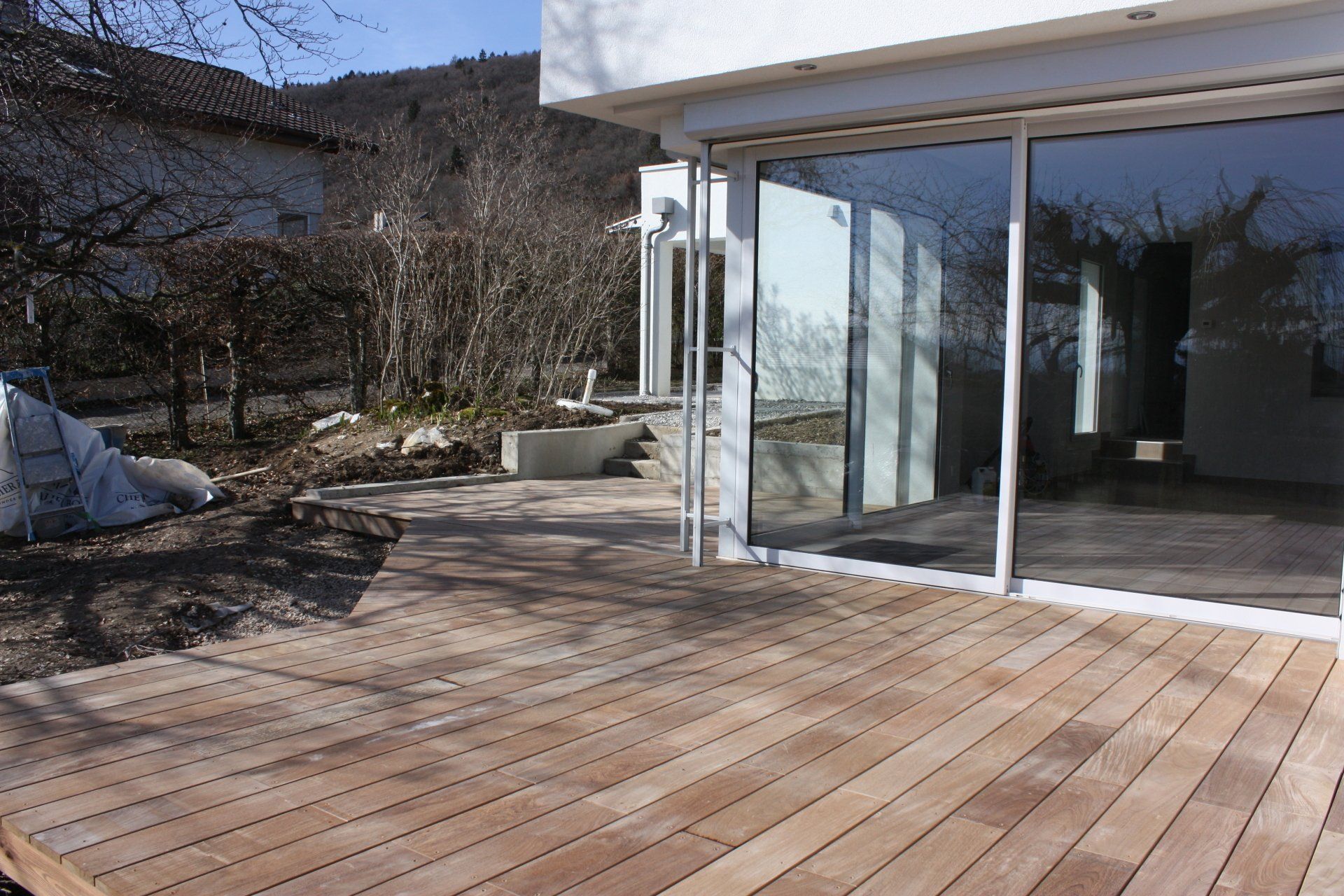 Terrasse en bois avec portes coulissantes en verre, bâtiment blanc et marches menant à une cour avec des arbres dénudés.