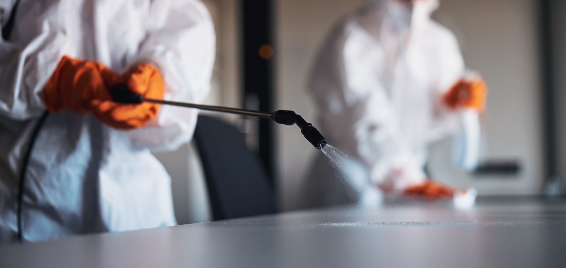 Deux personnes en combinaisons de protection blanches vaporisent du désinfectant sur une table.