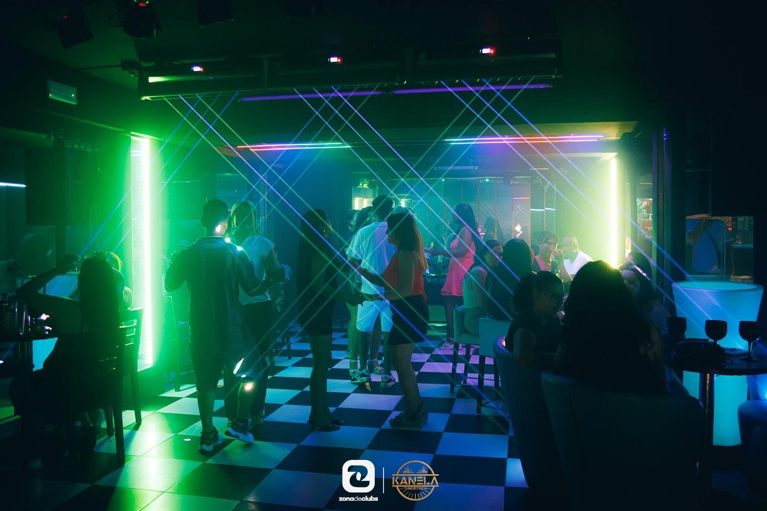 Gente bailando en un club iluminado con luces de neón verdes y moradas y un suelo a cuadros.