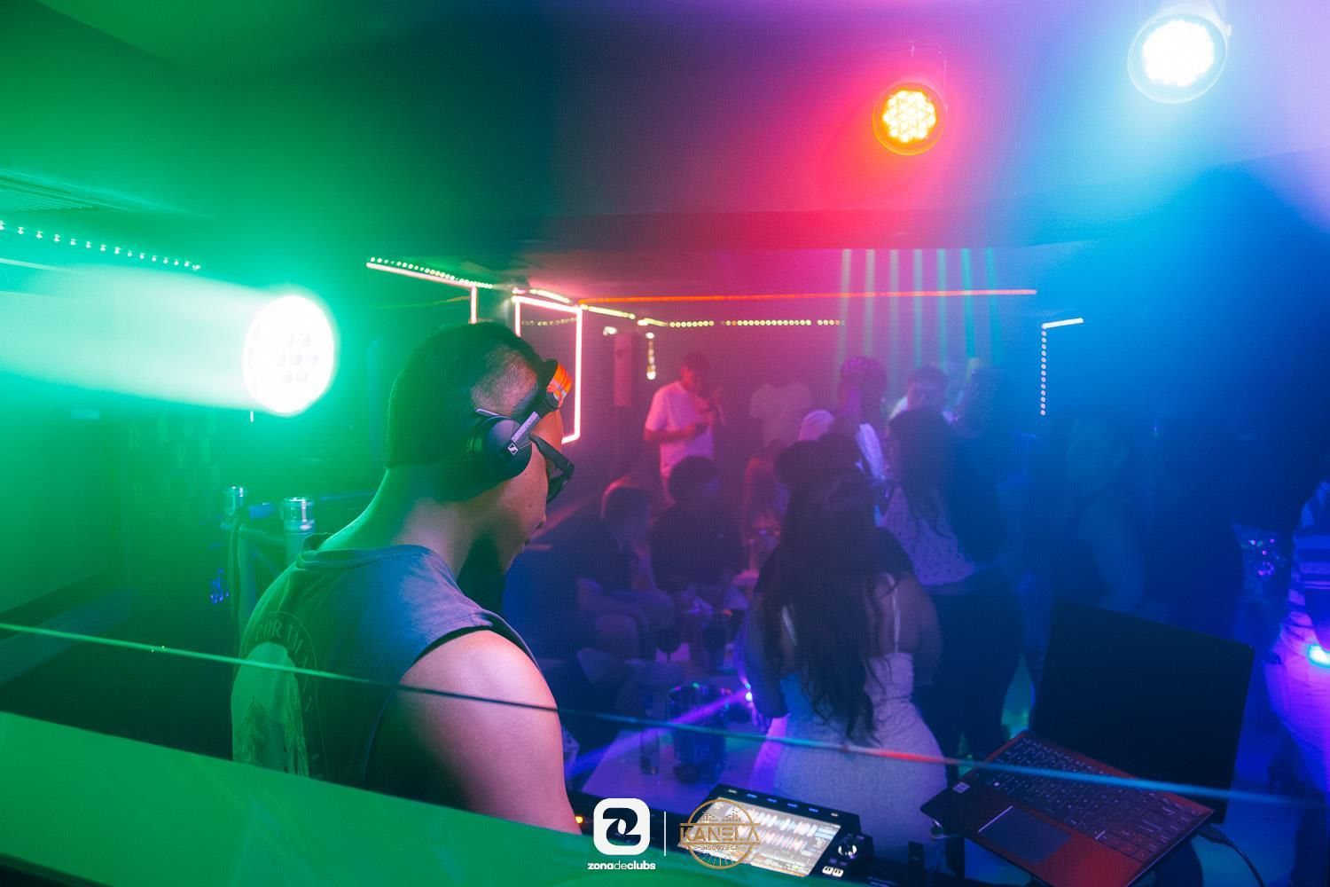 DJ en una discoteca iluminada con luces de neón de colores verde, rojo y azul sobre una multitud.