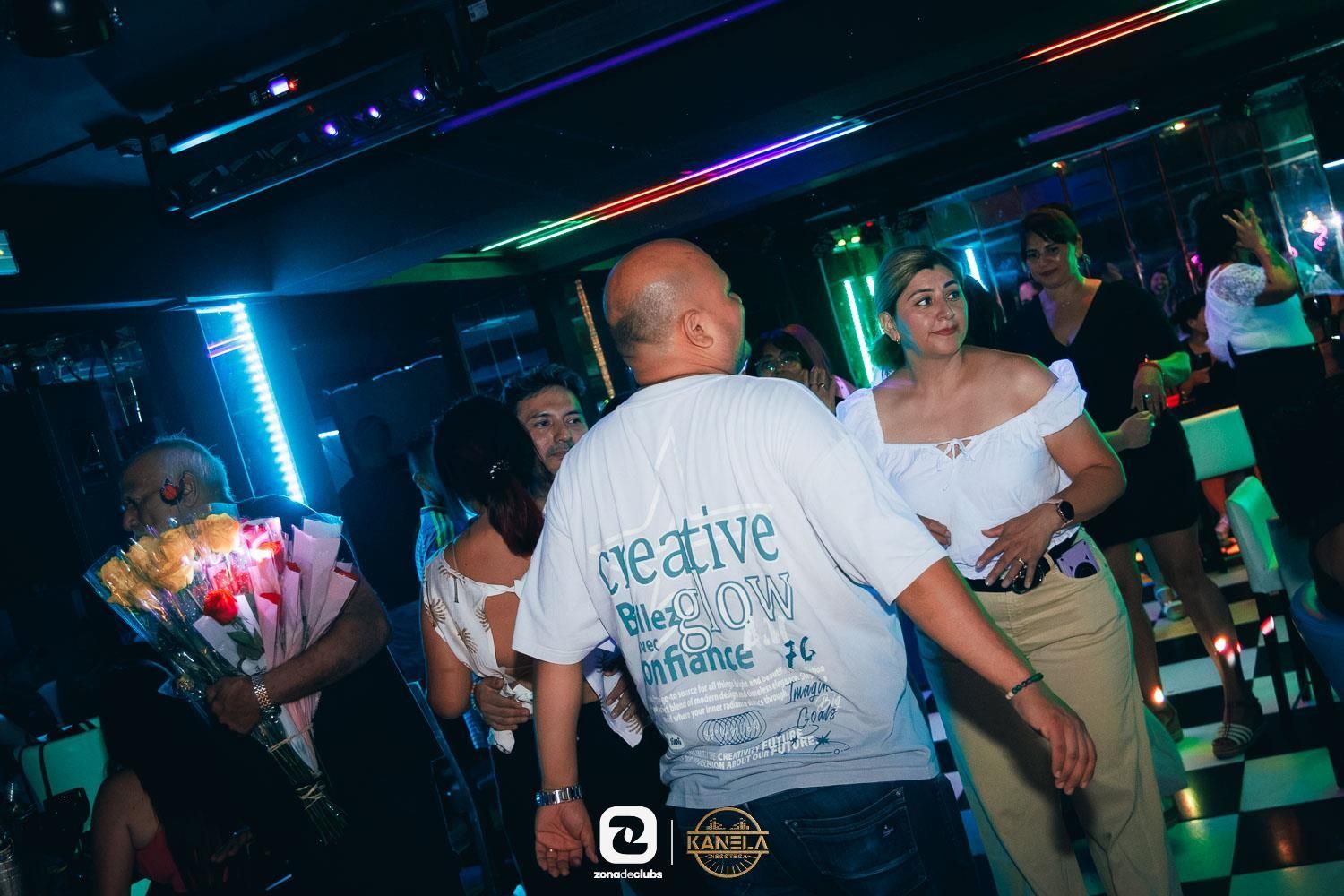 Gente bailando en una discoteca con poca luz, luces de neón de colores y un hombre con una camisa blanca en primer plano.