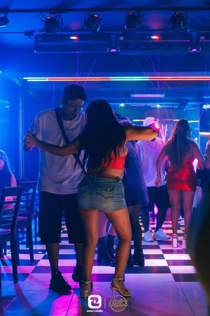 Gente bailando en un club iluminado con luces de neón y con un suelo de baldosas a cuadros resplandecientes.