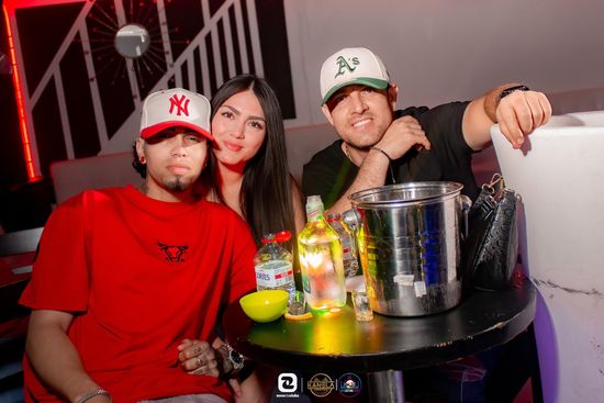 Tres personas posando en una mesa de discoteca con bebidas y una cubitera, vestidas con atuendos rojos y blancos.