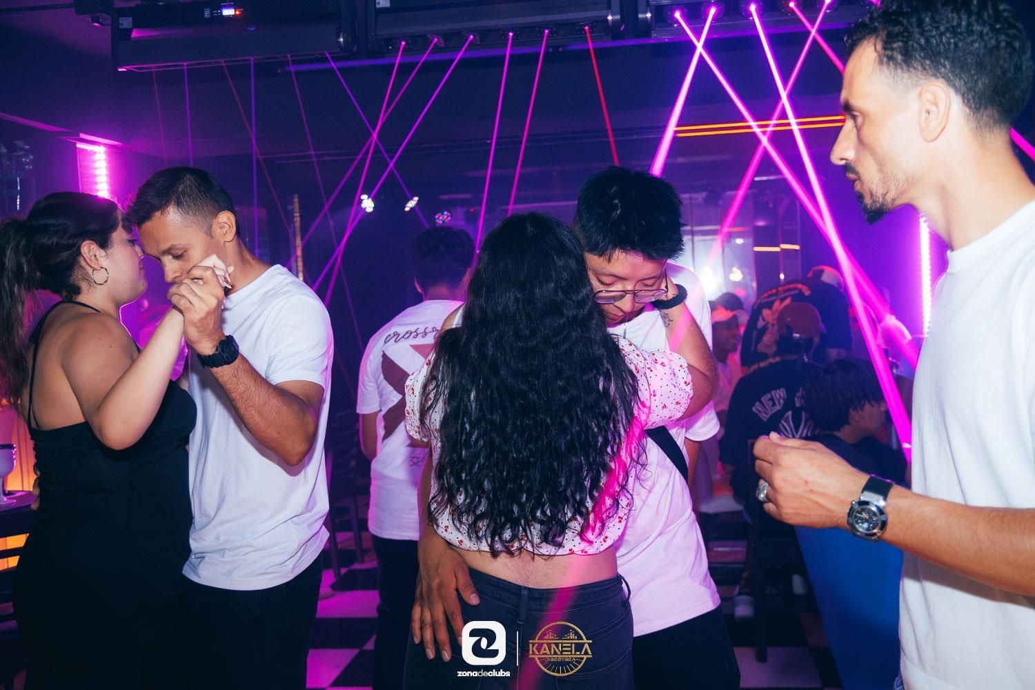 Gente bailando y charlando en un club iluminado con luces de neón moradas.