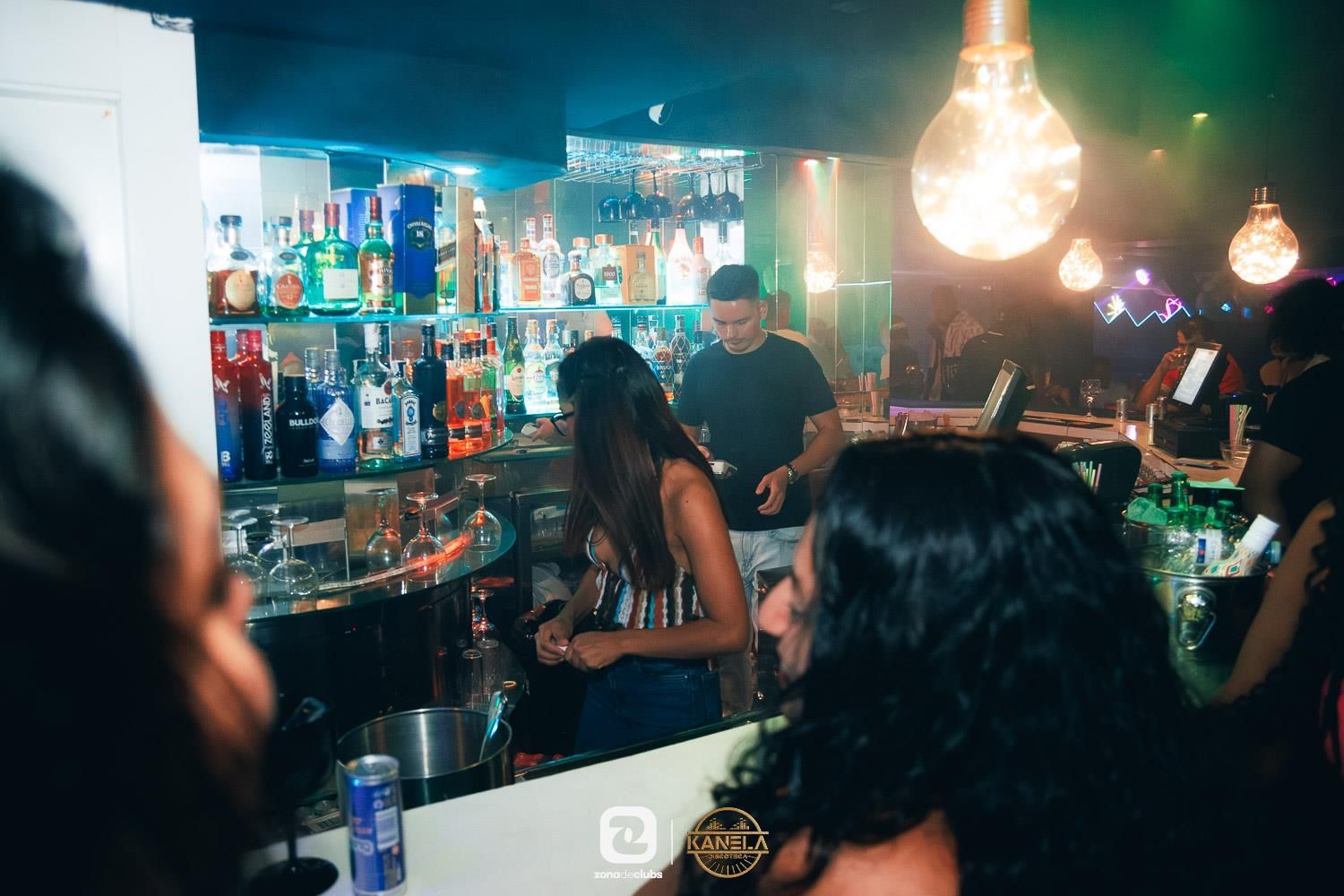Bar con iluminación tenue, gente charlando en la barra, botellas coloridas y lámparas colgantes.