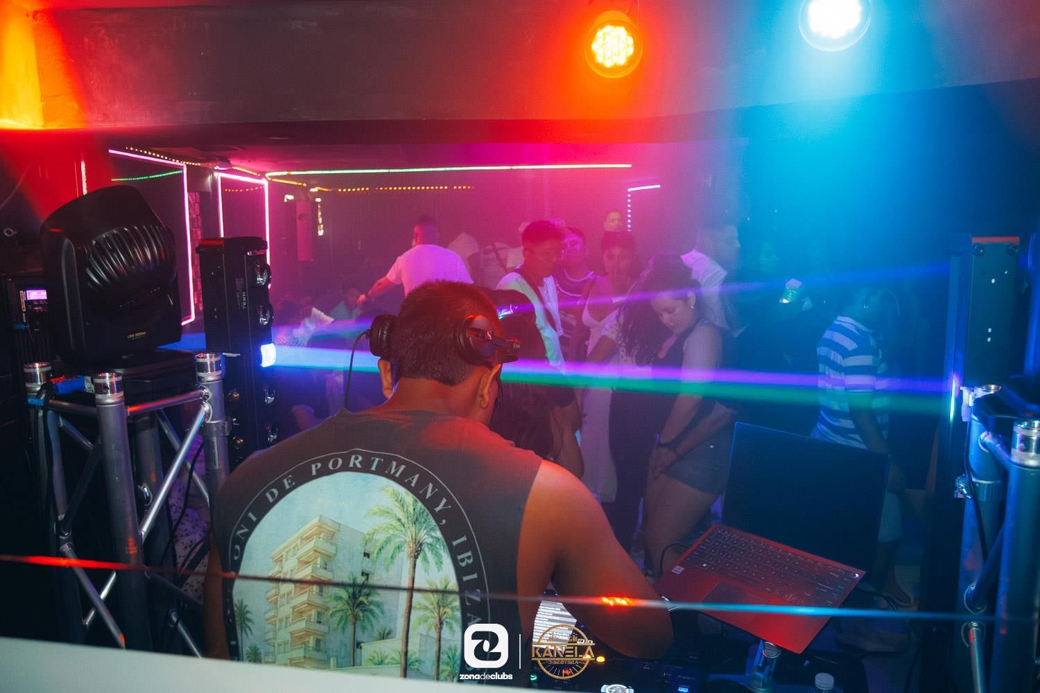 Cabina de DJ con gente bailando en una discoteca iluminada con luces de neón, con luces azules y rojas en lo alto.
