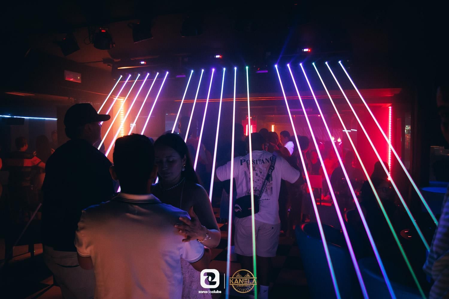 Gente bailando en un club oscuro con coloridos rayos láser atravesando la sala.
