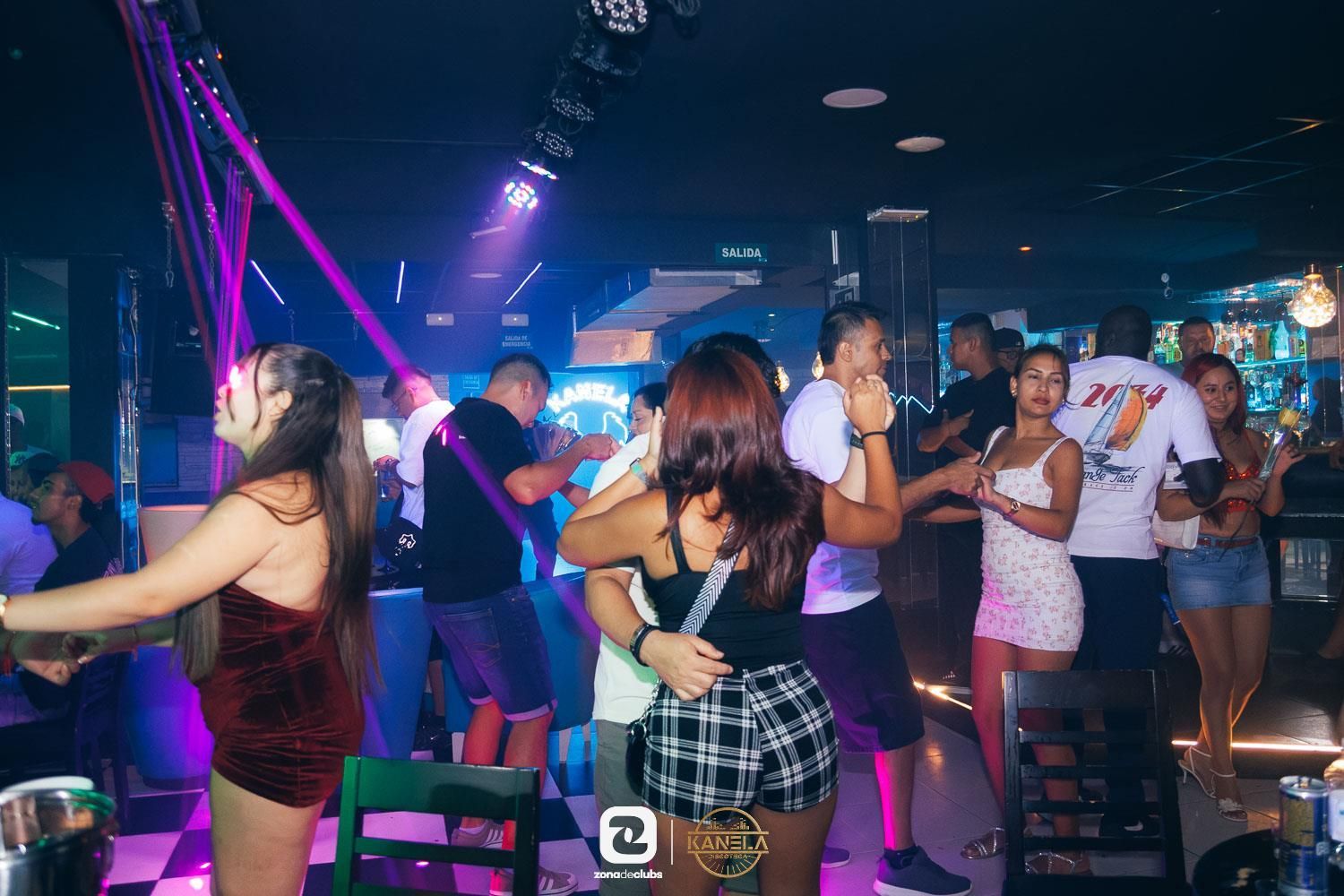 Gente bailando y socializando en una discoteca iluminada con luces de neón moradas y azules.