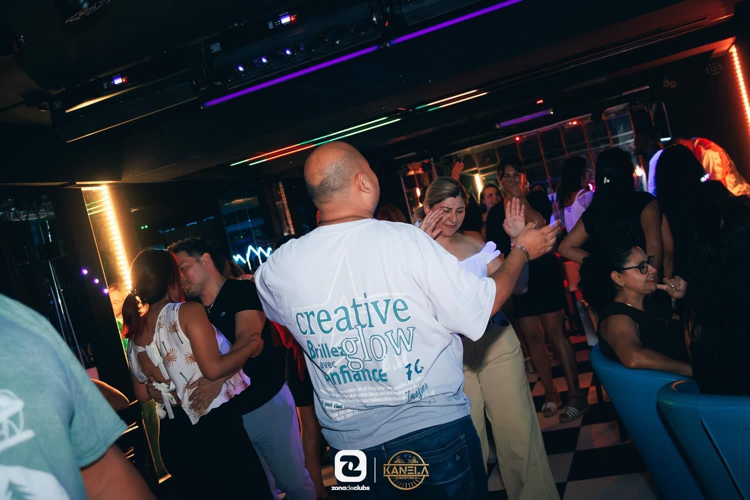 Discoteca abarrotada de gente bailando bajo luces de neón, vista desde detrás de un hombre con camisa blanca.