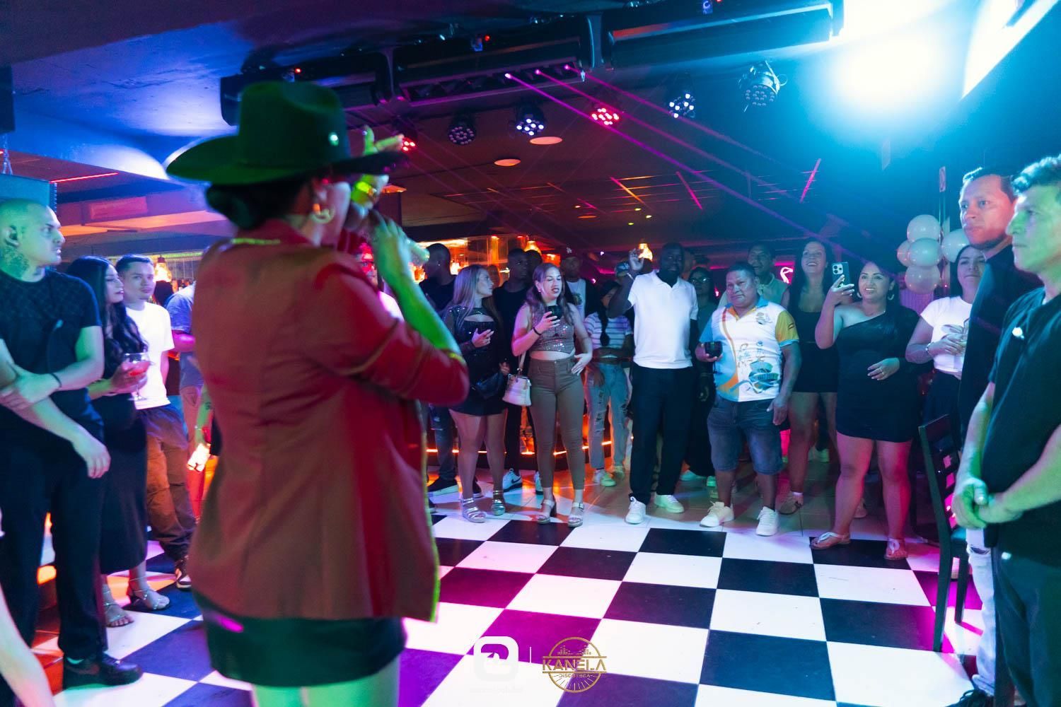 Una persona con sombrero verde y chaqueta roja canta en una pista de baile en blanco y negro en una discoteca, con una multitud observándola.