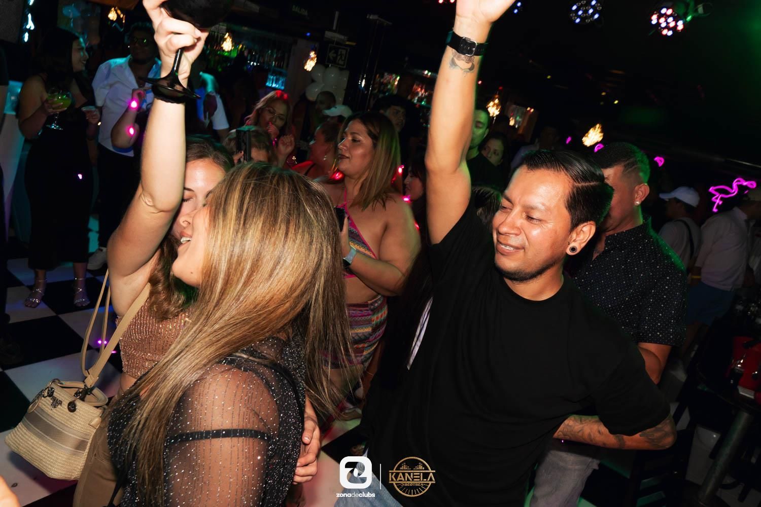 Gente bailando con los brazos en alto en una discoteca con poca luz, con luces de colores y bebidas sobre las mesas.
