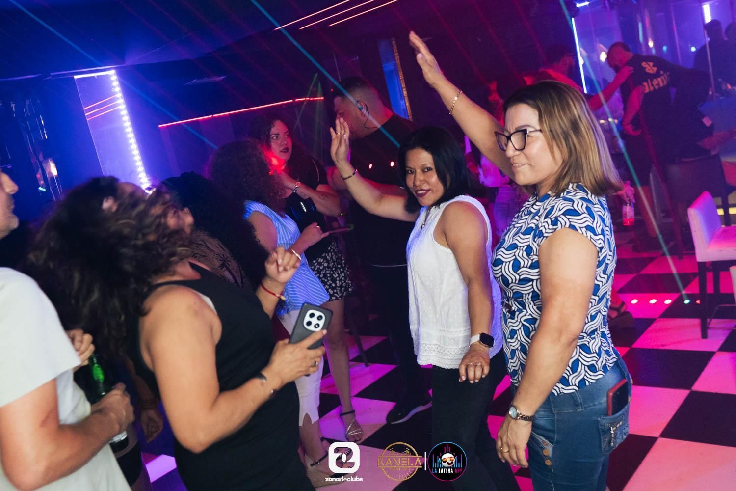 Gente bailando en una discoteca iluminada con luces de neón, con los brazos en alto y un escenario al fondo.