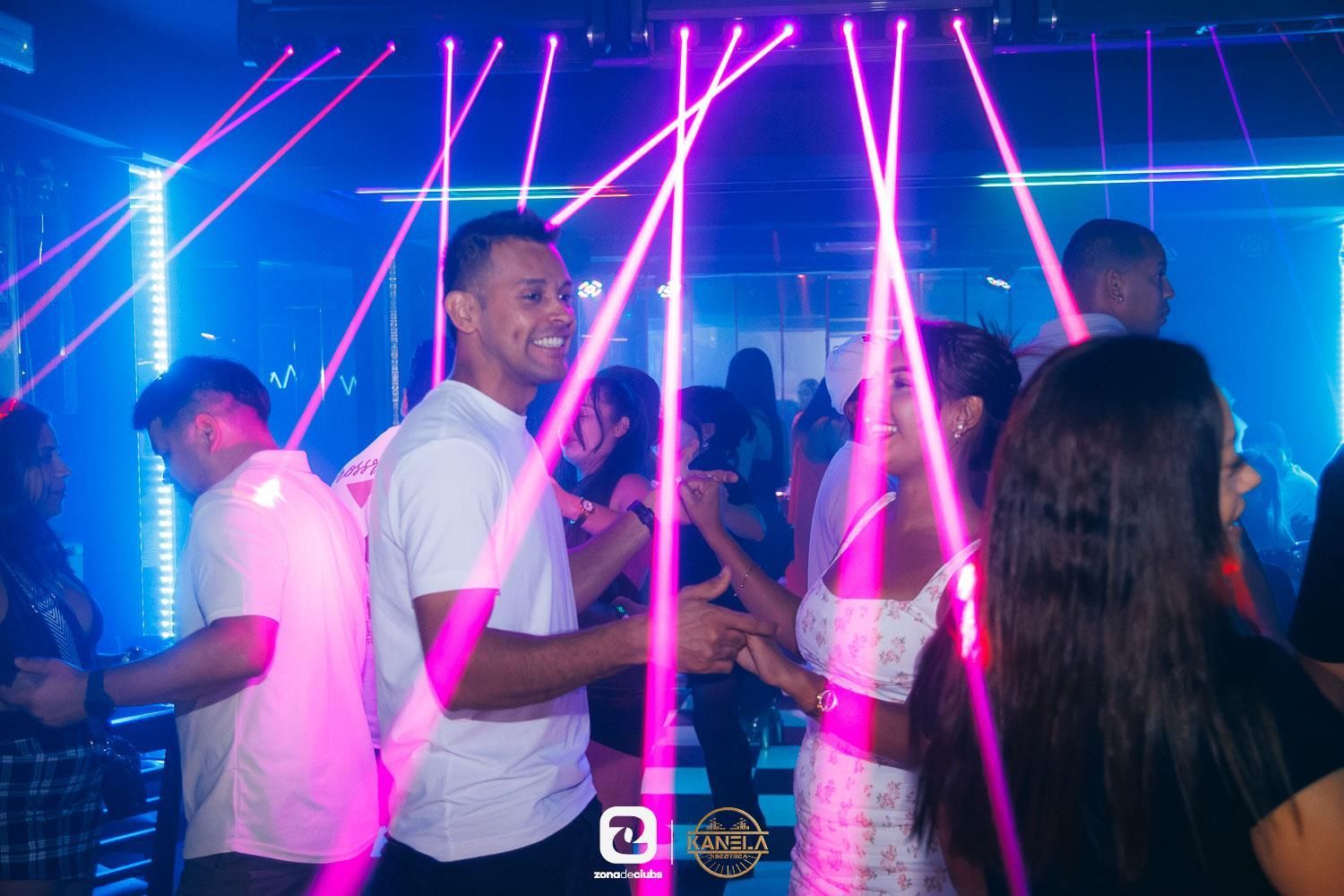 Discoteca abarrotada con luces láser rosas y gente bailando y hablando.