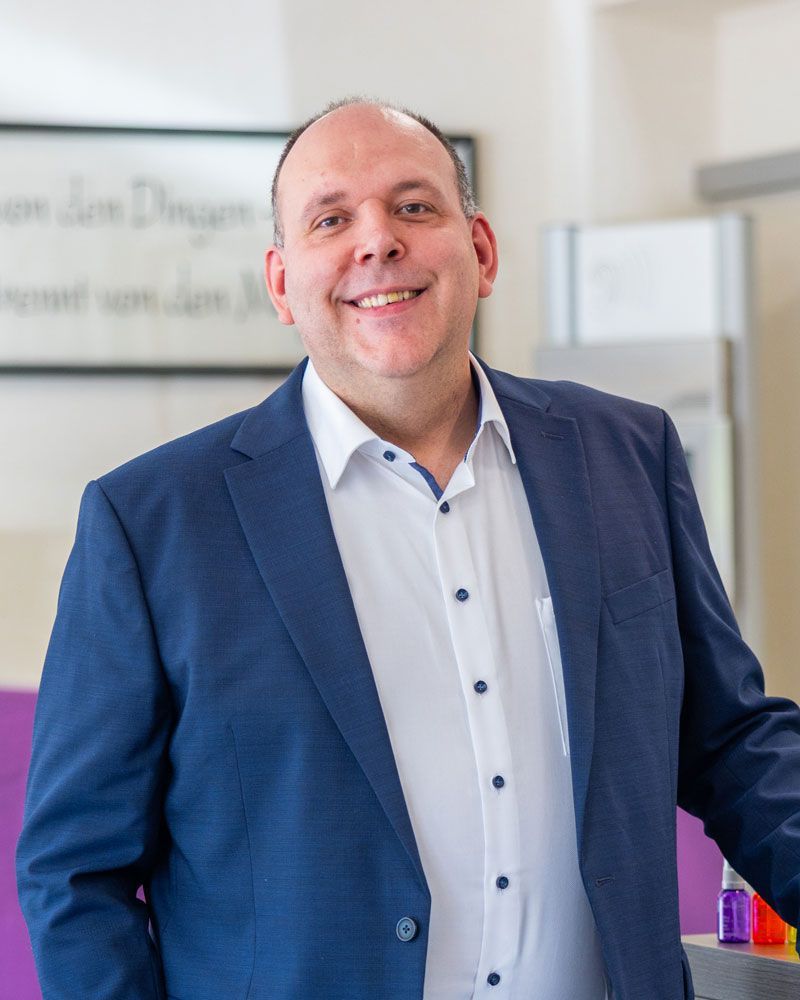 Mann mit Glatze, blauem Blazer und weißem Hemd, lächelt in die Kamera.