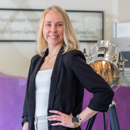 Eine Frau mit blonden Haaren und blauen Augen lächelt und trägt eine schwarze Jacke und eine Halskette .