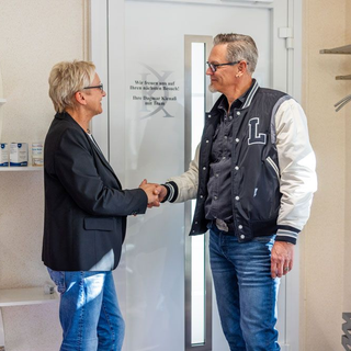 Zwei Personen geben sich in einem Büro die Hand. Mann in Collegejacke und Jeans, Frau in Blazer und Jeans.