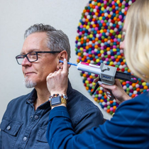 Eine Frau untersucht mit einem spritzenähnlichen Instrument das Ohr eines Mannes. Sie befinden sich im Ohr, im Hintergrund ist ein farbenfrohes Kunstwerk zu sehen.