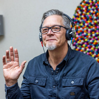Mann mit ergrautem Haar und Brille winkt und trägt Kopfhörer. Bunte Kunst im Hintergrund.