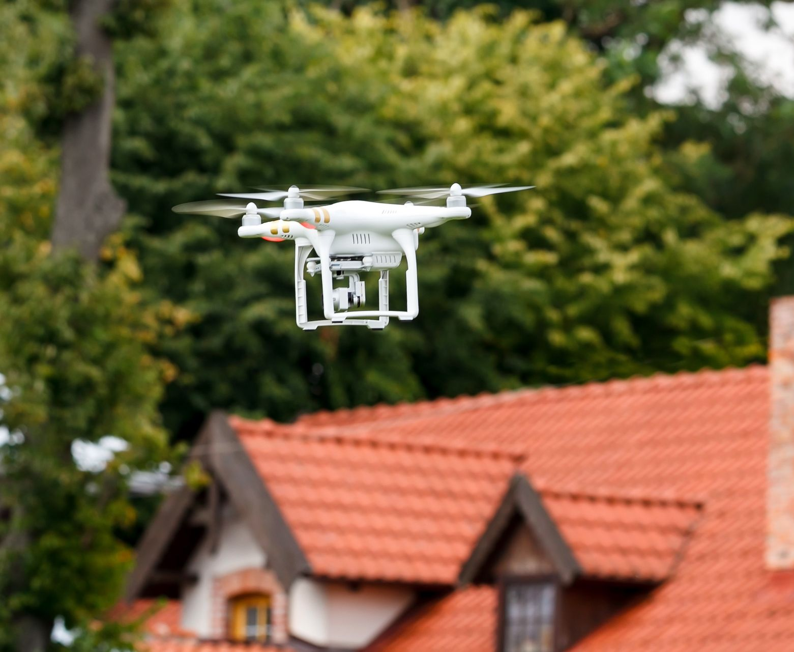 Drone blanc planant devant une maison avec un toit de tuiles orange et des arbres verts.