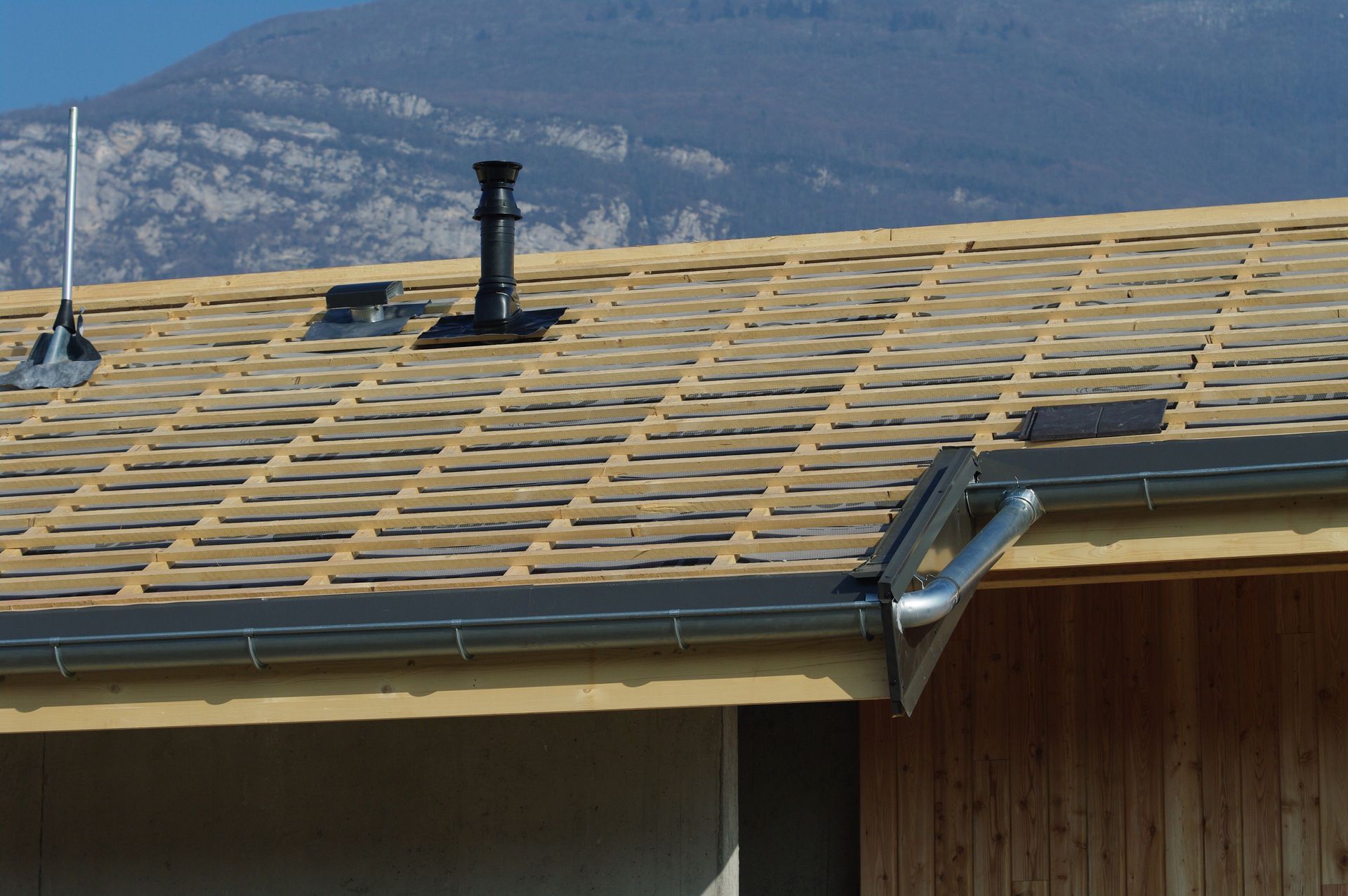 Toiture en construction avec poutres en bois, ventilation noire et gouttière, sur fond de montagne.