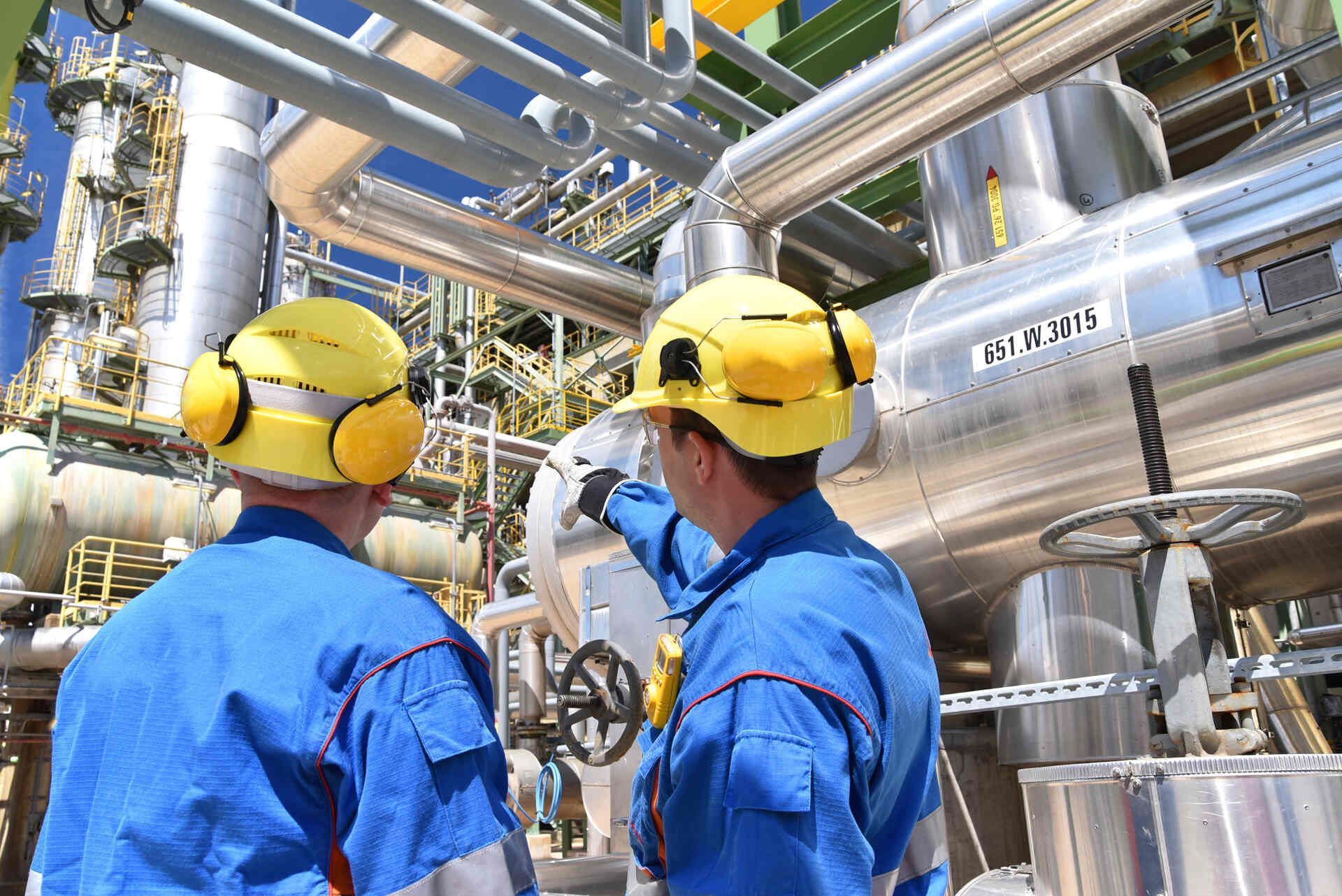 Zwei Männer mit Schutzhelmen und Ohrenschützern arbeiten in einer Fabrik.