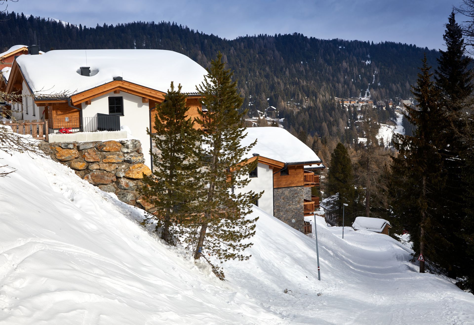 Verschneite Almhütten eingebettet in einen schneebedeckten Hang, Bäume im Vordergrund, Wald und Berge im Hintergrund.