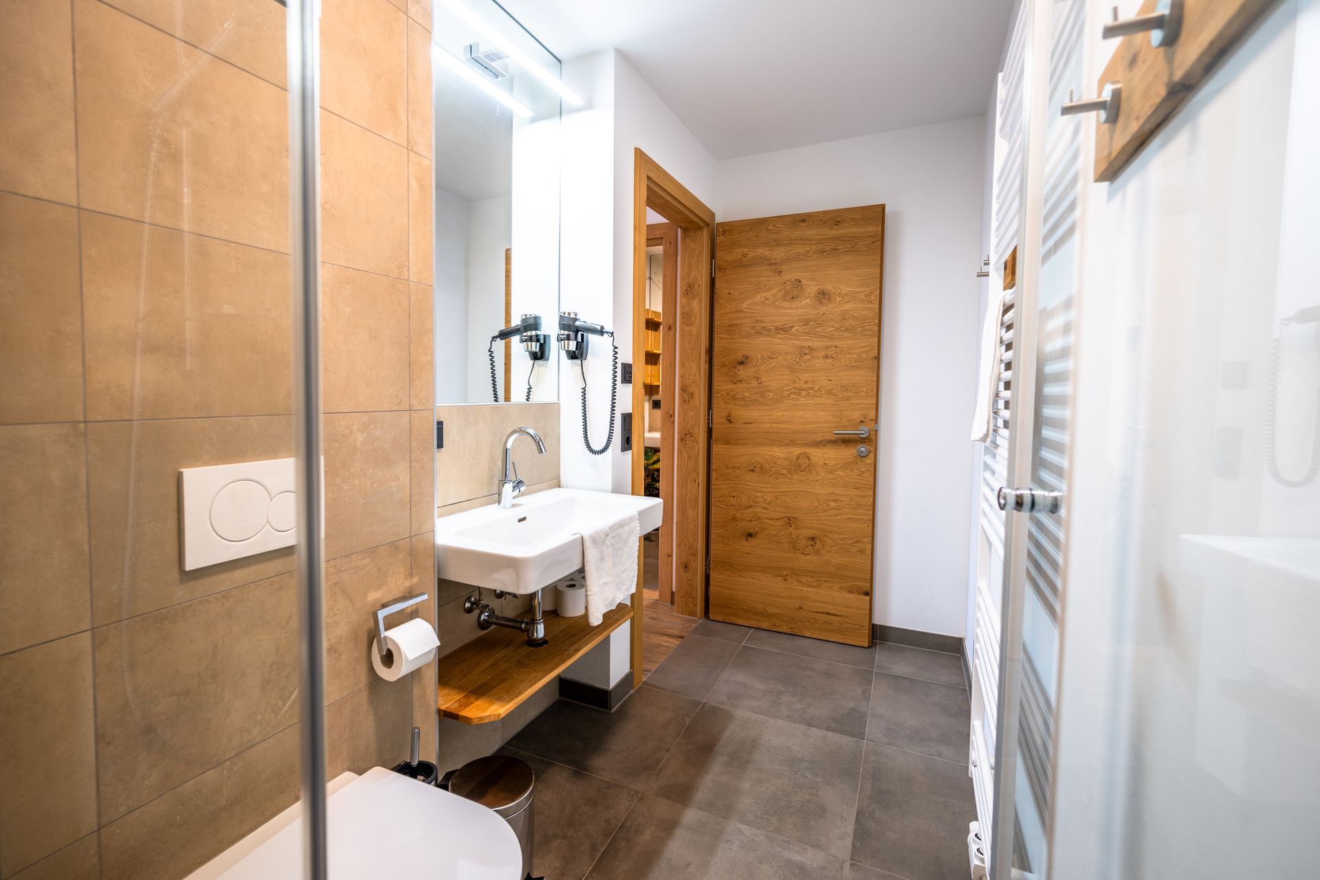 Badezimmer mit Dusche, Waschbecken, Toilette, Holztür und grauem Fliesenboden.