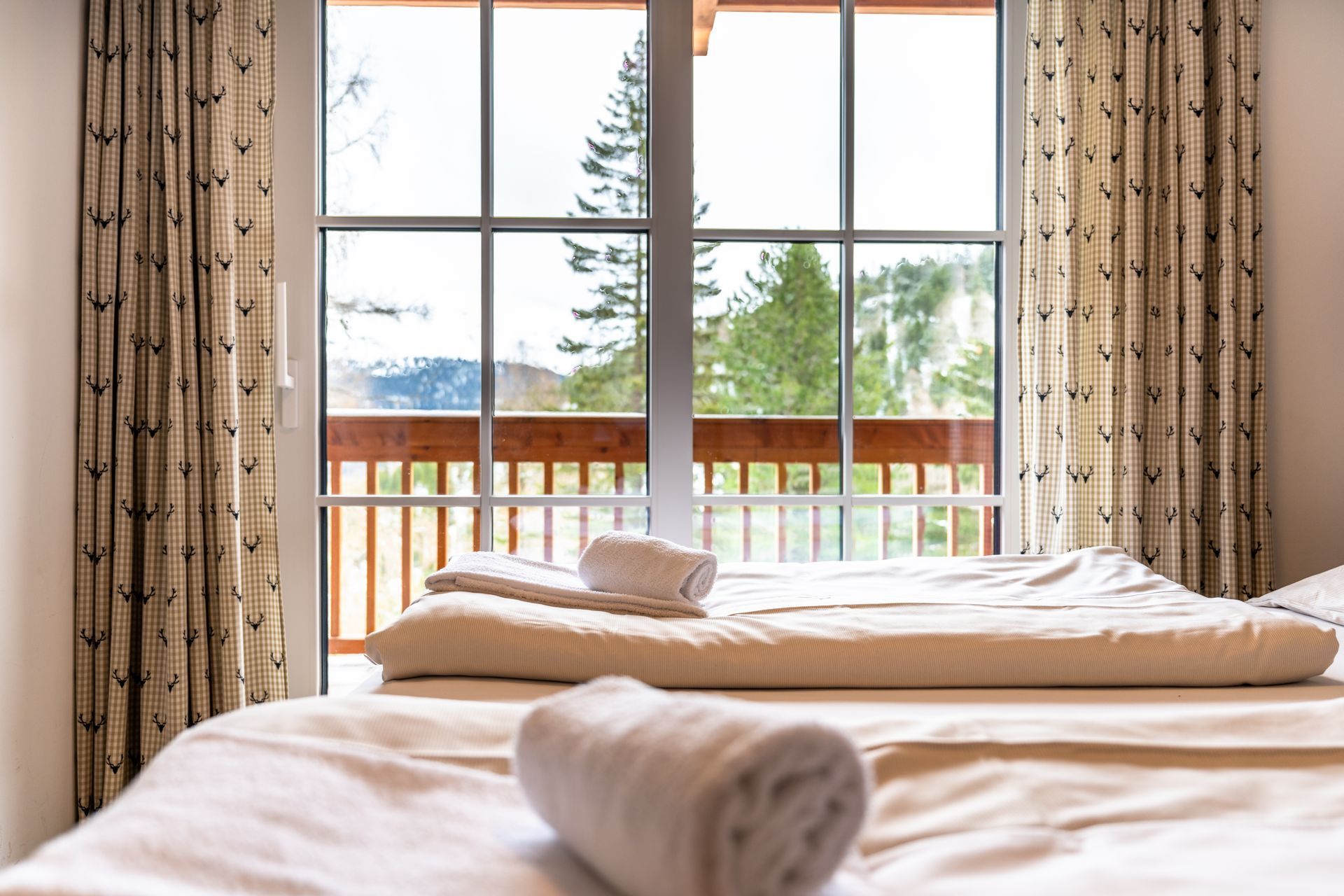 Schlafzimmer mit zwei Betten, Handtüchern und einem Fenster mit Blick auf einen Balkon und Bäume.
