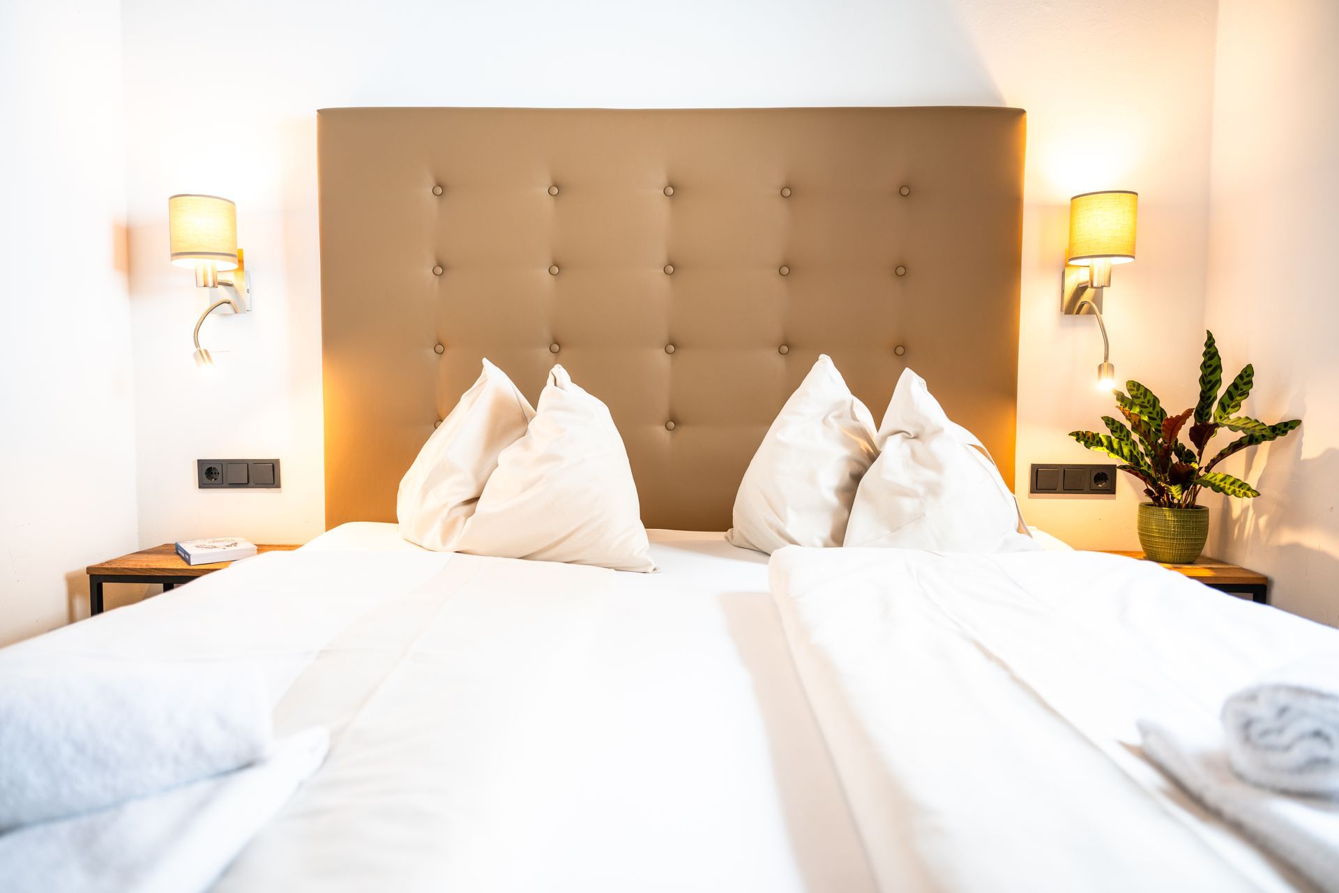 Schlafzimmer mit weißer Bettwäsche, hellbraunem Kopfteil und Wandlampen.