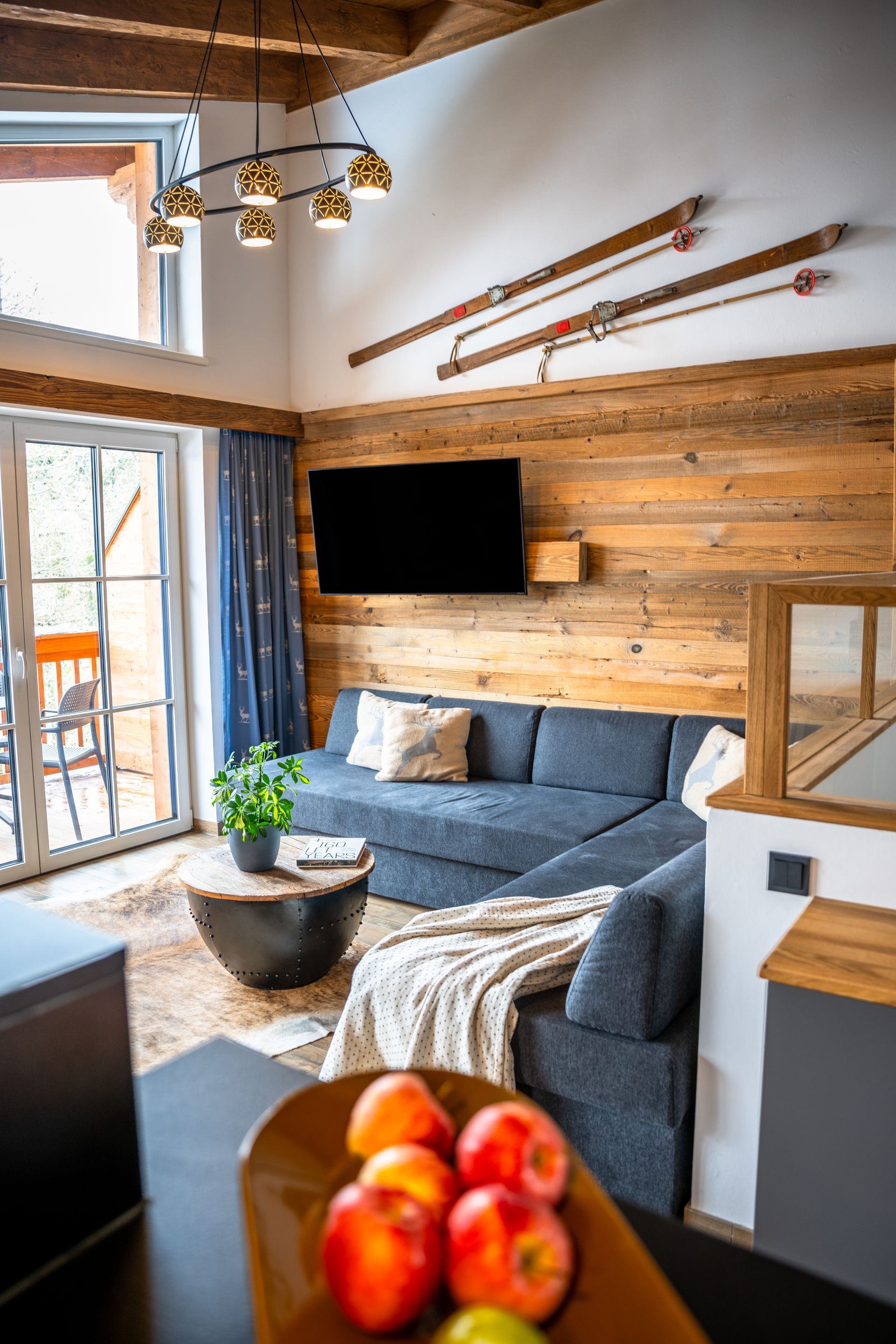 Gemütliches Wohnzimmer mit holzgetäfelter Wand, blauem Sofa und Skiern. Mit Fernseher, Couchtisch und Obstschale.