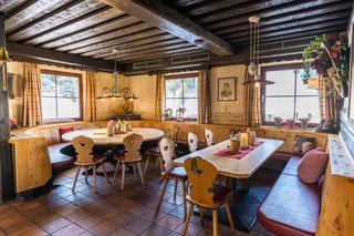 Gemütlicher, rustikaler Speisesaal mit Holztischen, Stühlen und Bänken. Fenster sorgen für natürliches Licht, Ziegelboden.