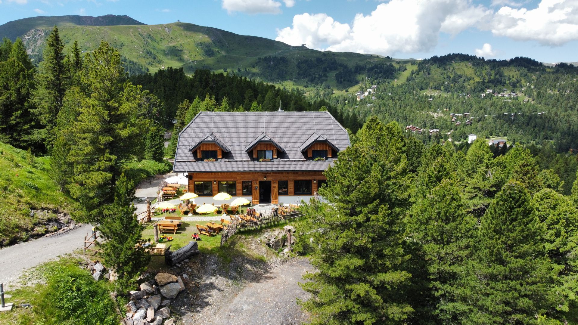 Holzgebäude mit Sitzgelegenheiten im Freien, eingebettet in eine Berglandschaft mit Bäumen und bewölktem Himmel.