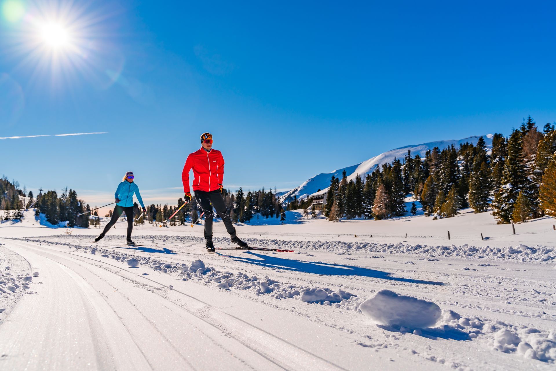Zwei Personen laufen auf Skiern auf einer verschneiten Loipe unter strahlender Sonne, im Hintergrund sind Berge und Bäume zu sehen.