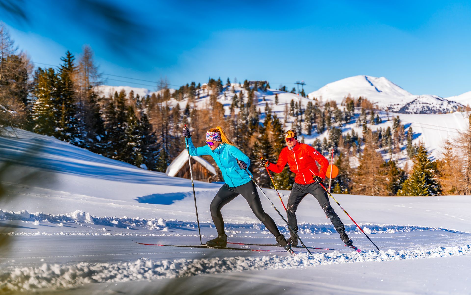 Zwei Personen beim Langlaufen auf einer verschneiten Loipe in den Bergen, strahlend blauer Himmel.