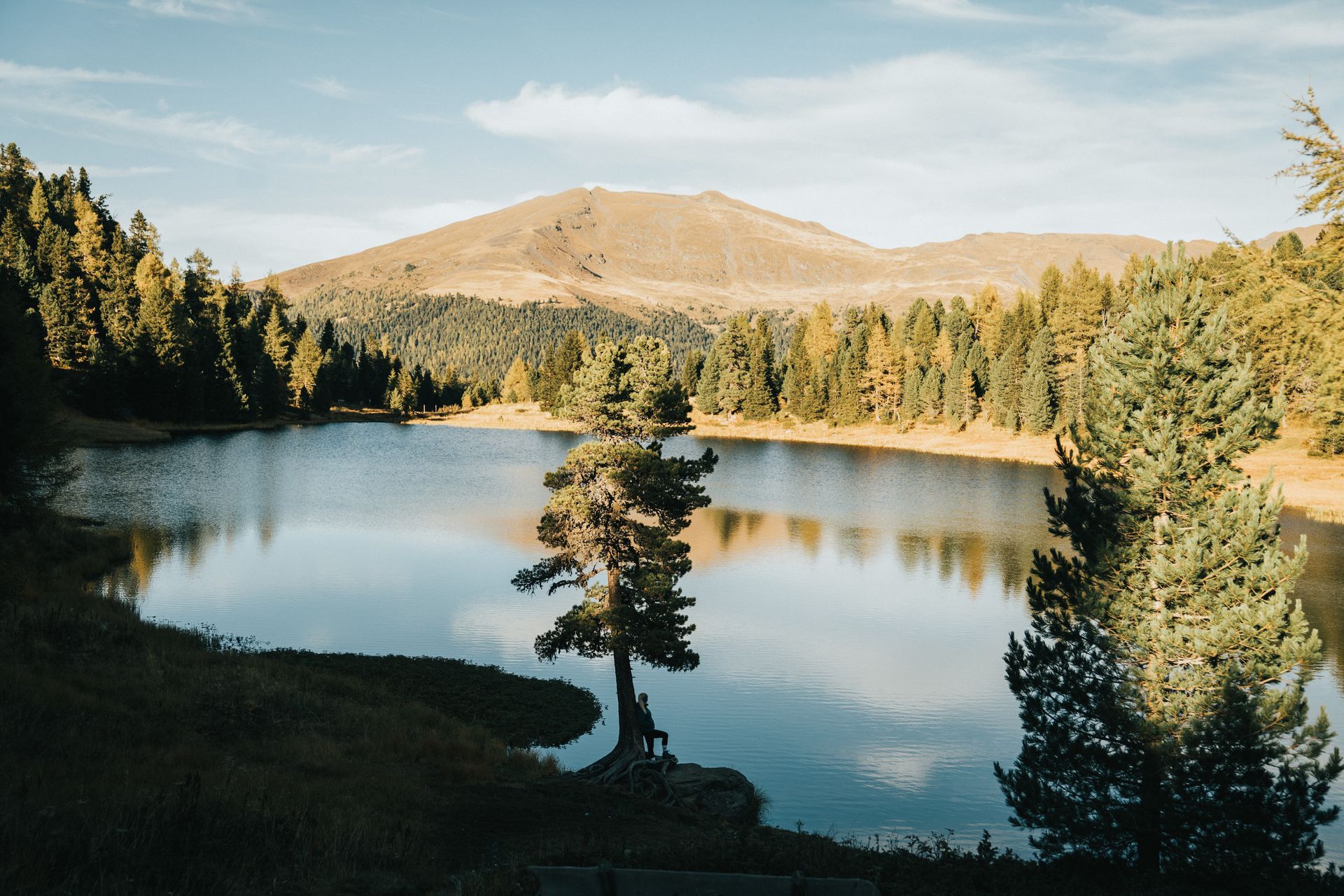 See umgeben von grünen Bäumen und einem Berg unter einem sonnigen Himmel.