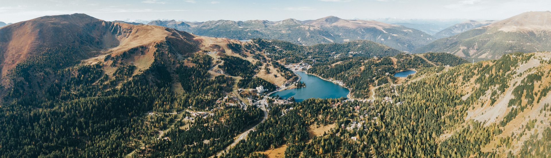 Luftaufnahme einer Bergkette mit einem blauen See, umgeben von grünen Bäumen und braunen Hügeln.