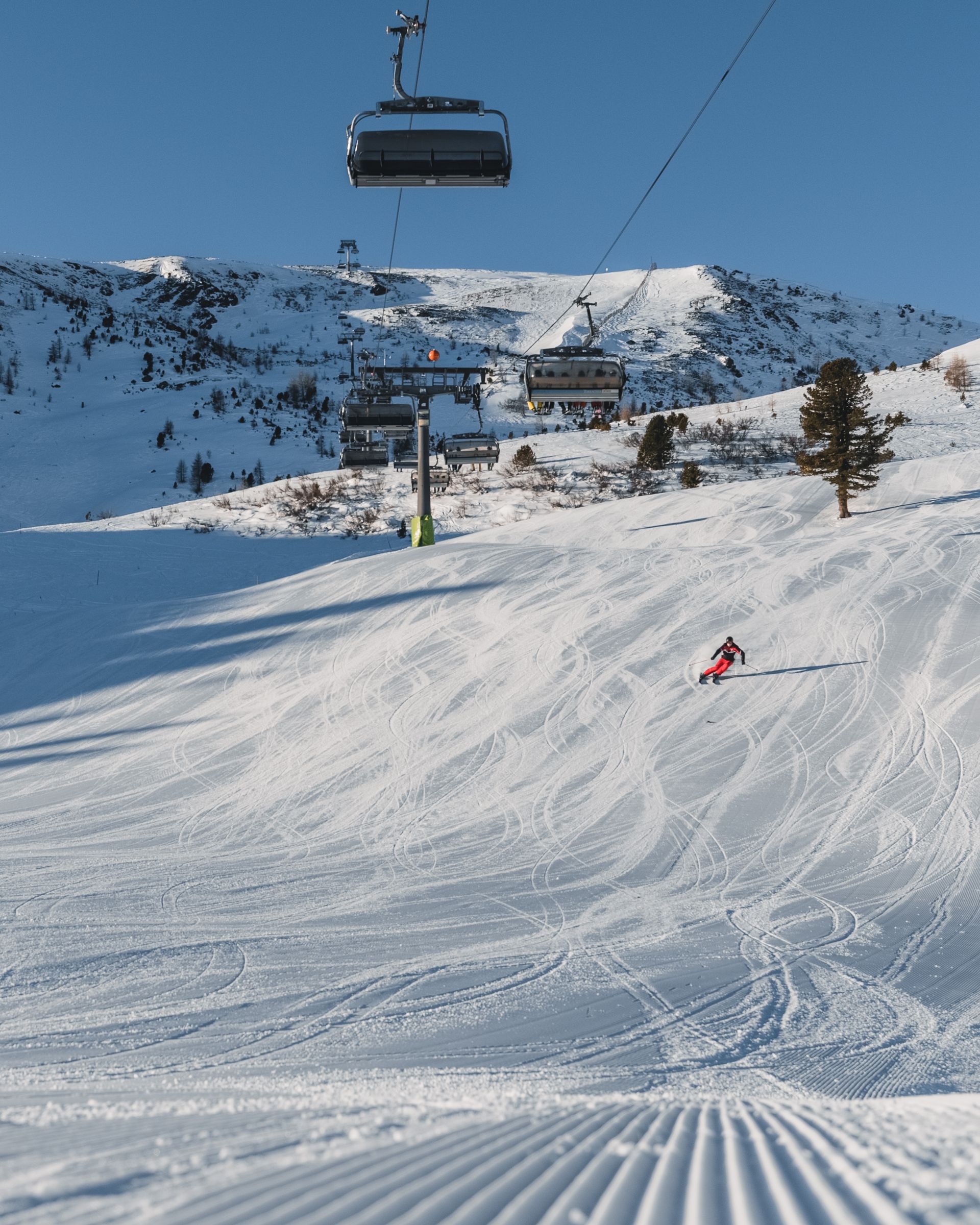 Skifahrer fährt einen schneebedeckten Hang hinunter, über ihm ein Skilift. Blauer Himmel, Berge im Hintergrund.
