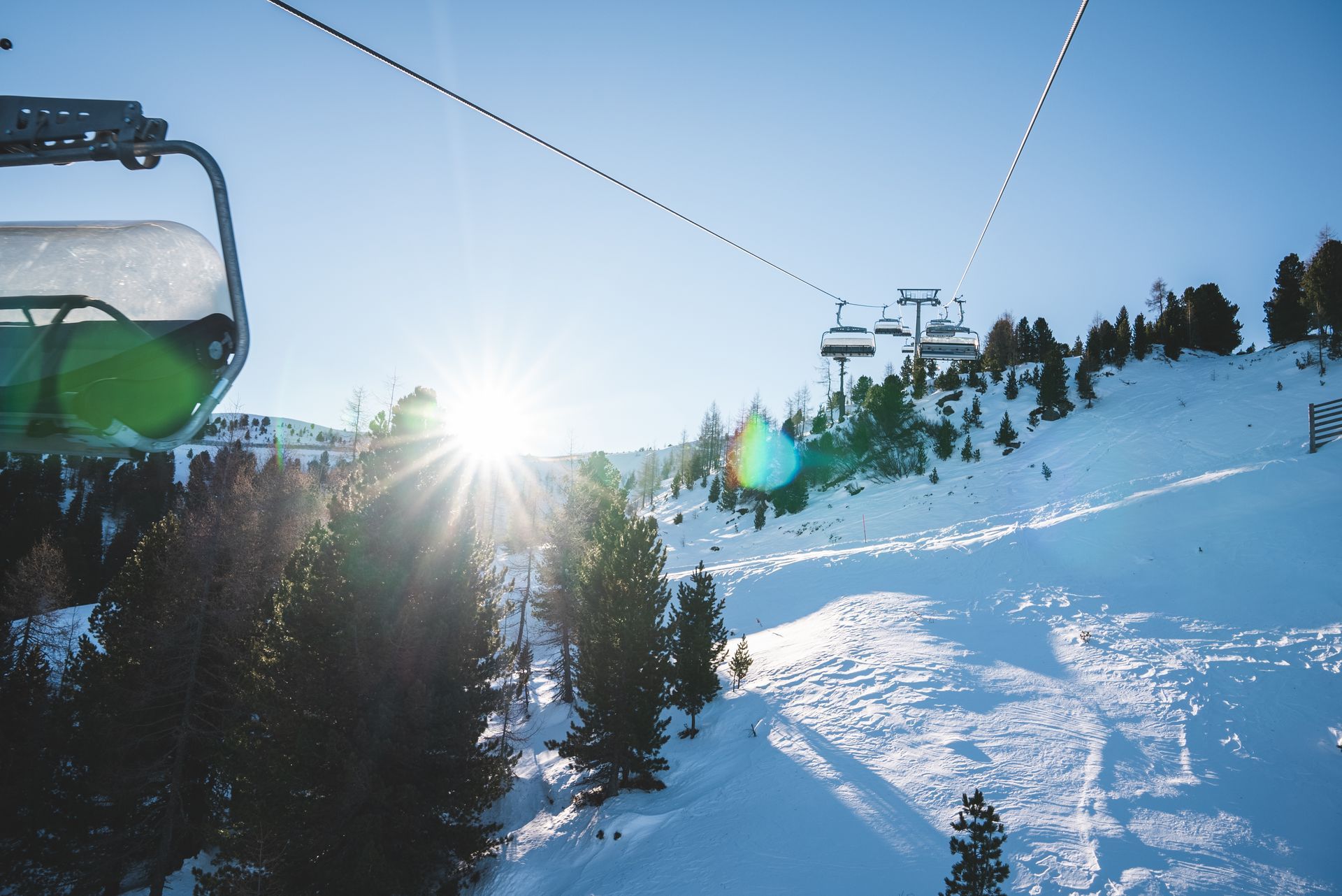 Skilift fährt einen verschneiten Berghang hinauf, im Hintergrund die strahlende Sonne.