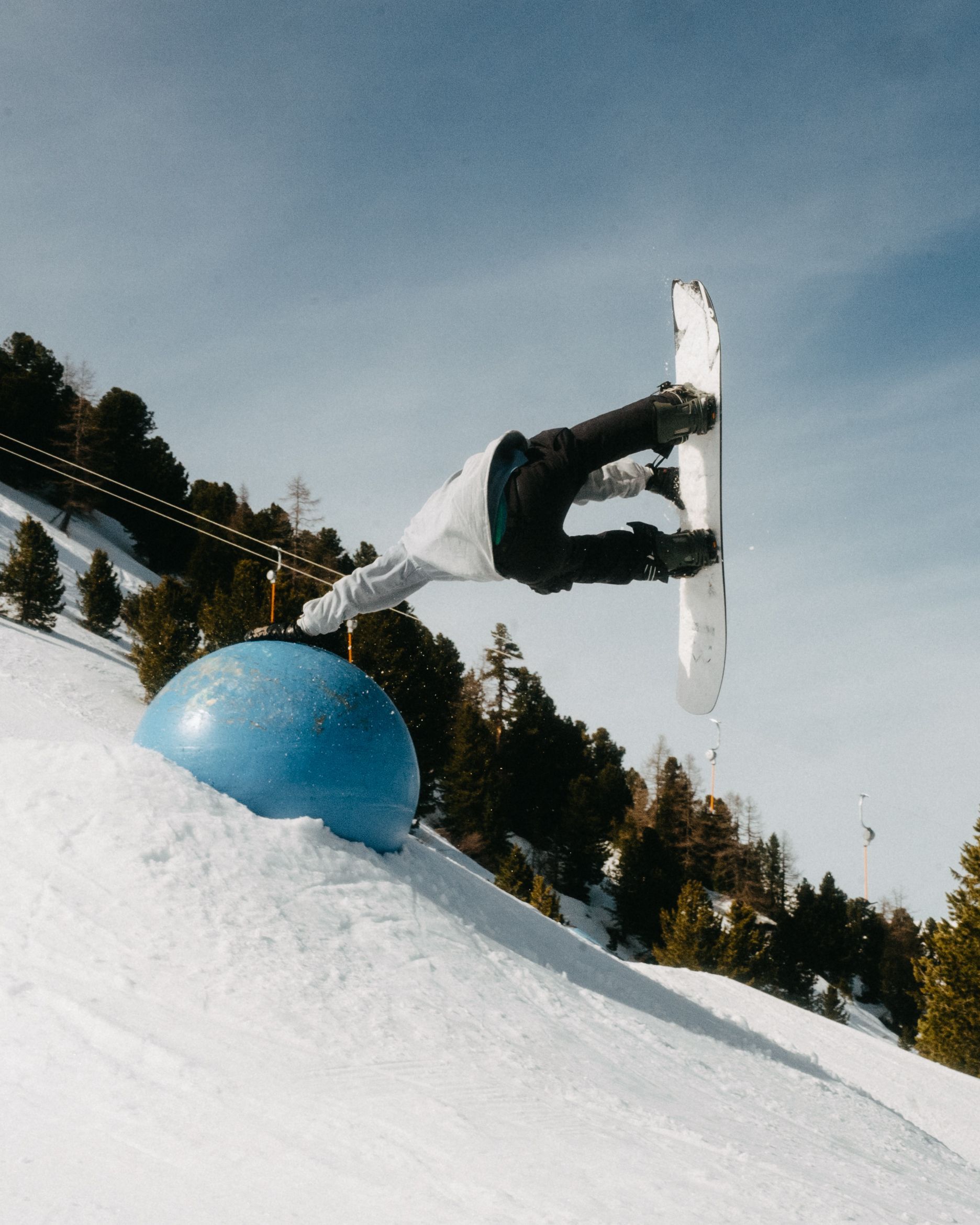 Snowboarder in der Luft über einer blauen Kugel auf einem schneebedeckten Hang unter einem sonnigen Himmel.