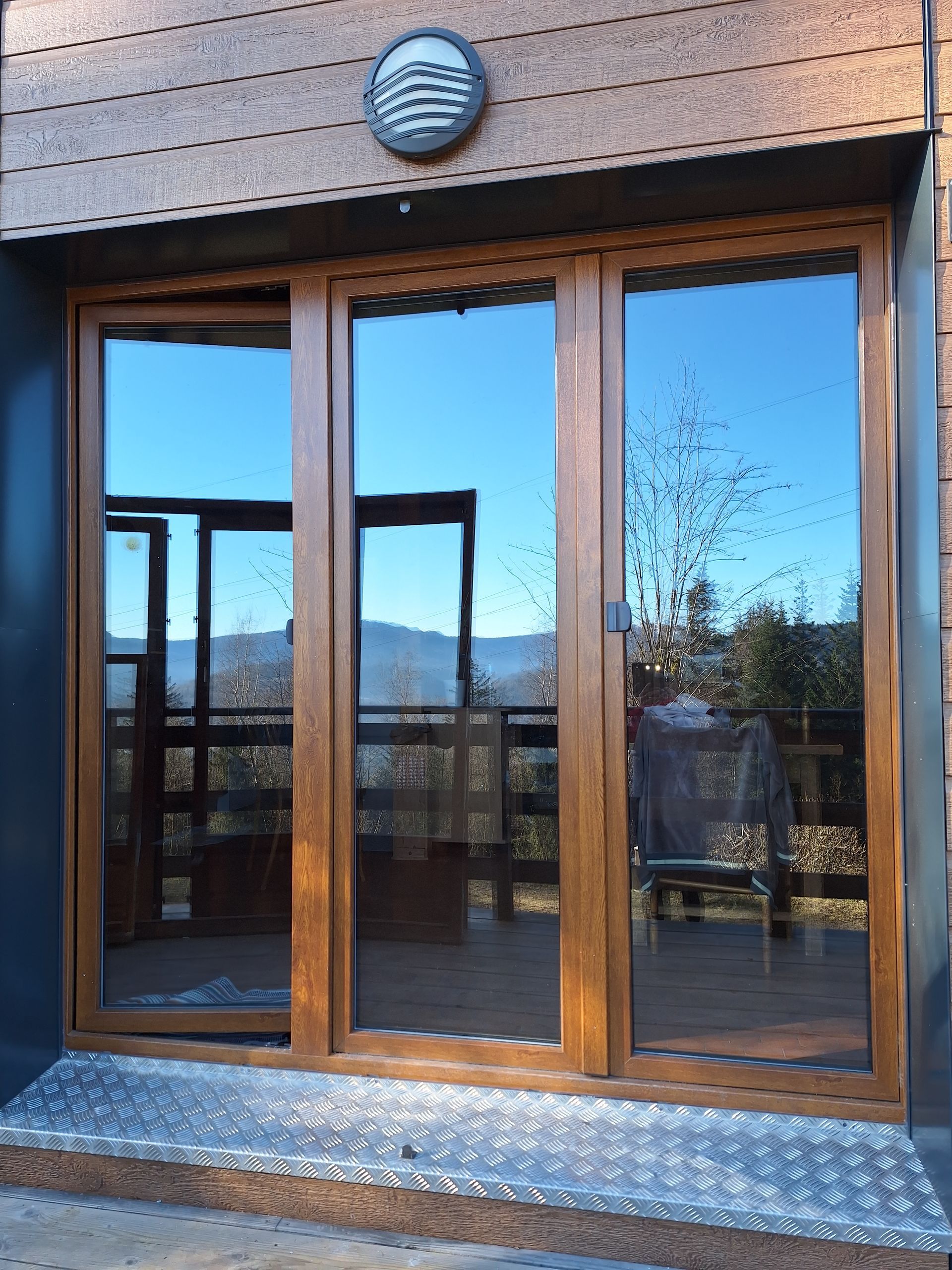 Des portes coulissantes en bois et verre à trois panneaux reflètent le ciel et une terrasse, devant un bâtiment lambrissé.