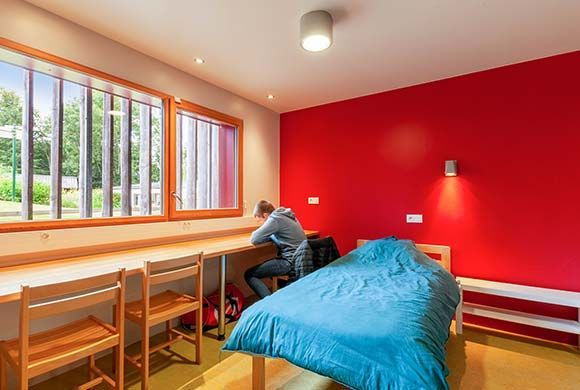 Une personne est assise à un bureau dans une pièce au mur rouge, regardant par la fenêtre. Un lit se trouve à sa droite.