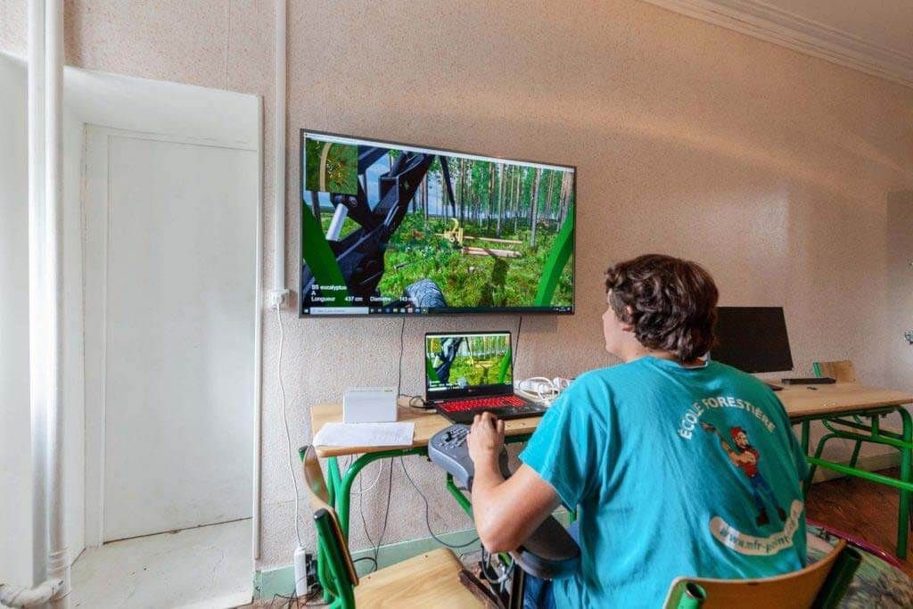 Personne assise, jouant à un jeu vidéo sur grand écran et ordinateur portable. Chemise bleu clair, murs beiges, bureau.