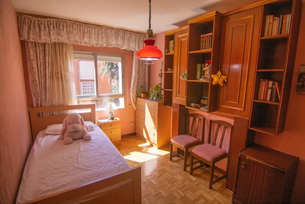 Habitación acogedora con cama individual, osito de peluche, armarios de madera y una ventana soleada con lámpara colgante roja.